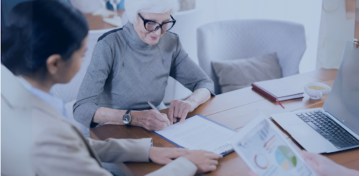 An elderly woman signs documents at a table, accompanied by two advisors. The setting includes a laptop, charts, and paperwork, indicating a professional consultation.