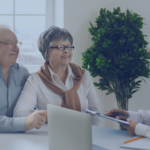 An elderly couple sitting across from a young female advisor in an office setting. The advisor is smiling and holding a clipboard, while the couple looks engaged and attentive.