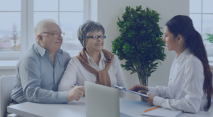 An elderly couple sitting across from a young female advisor in an office setting. The advisor is smiling and holding a clipboard, while the couple looks engaged and attentive.