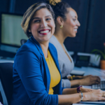 A group of professionals working at their desks with laptops. A woman in the foreground, dressed in a blue blazer and yellow top, is smiling at the camera.