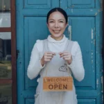 Smiling woman holding a “Welcome, We Are Open” sign in front of a blue door, with a plant and window displaying decor on the side.
