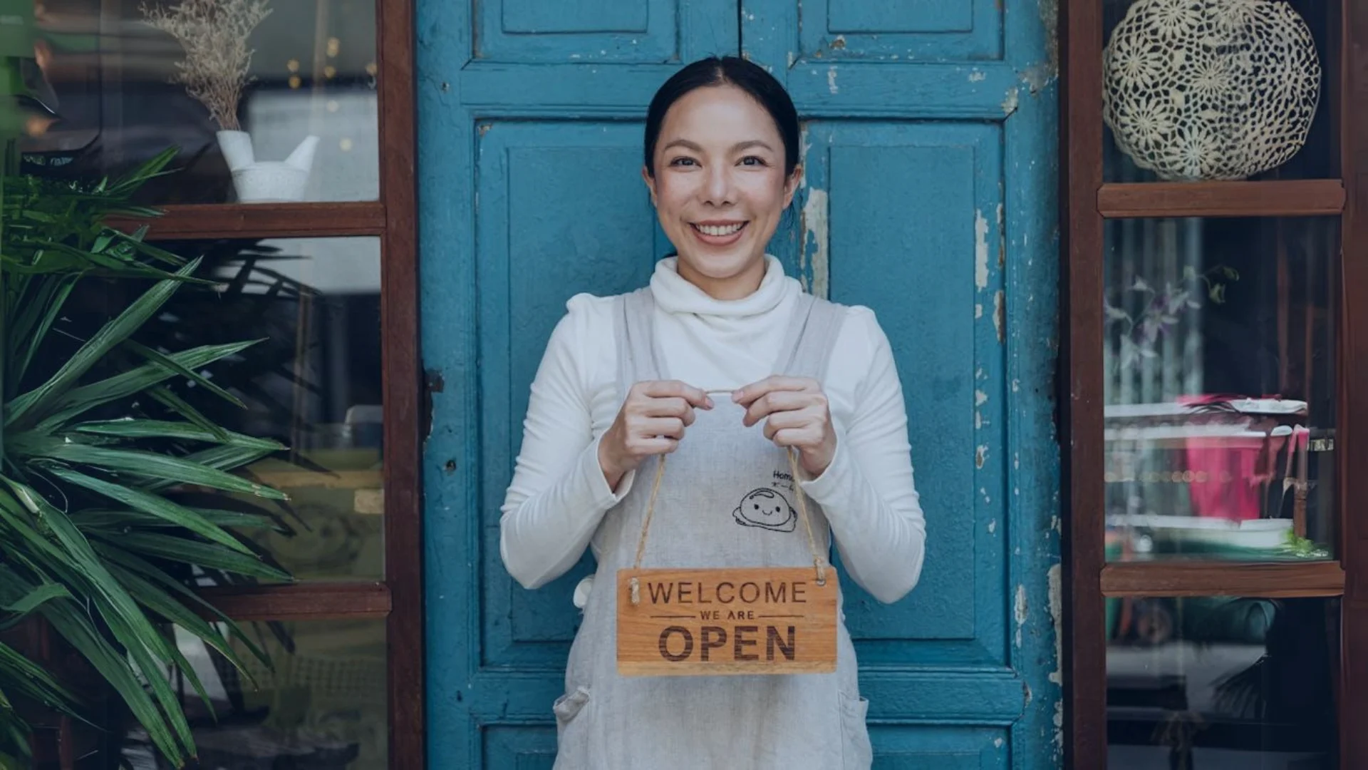 Smiling woman holding a “Welcome, We Are Open” sign in front of a blue door, with a plant and window displaying decor on the side.