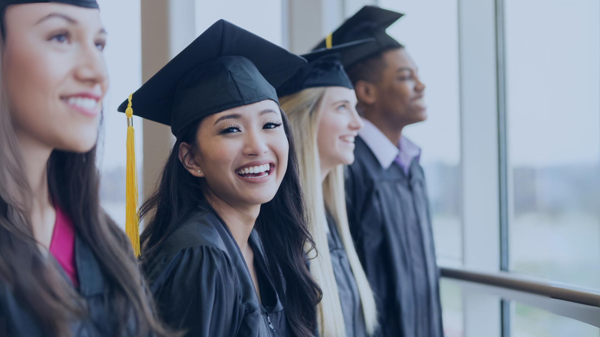 Group of smiling graduates in caps and gowns standing by a window.