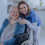 A smiling elderly woman in a wheelchair with a caregiver in blue scrubs posing together in a bright kitchen.