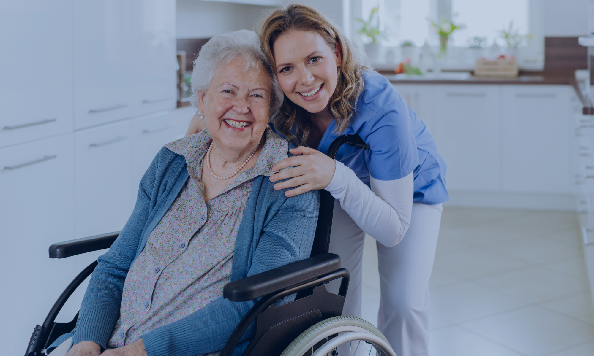 A smiling elderly woman in a wheelchair with a caregiver in blue scrubs posing together in a bright kitchen.