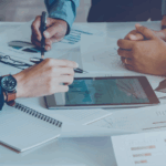 Business professionals analyzing financial data and performance metrics on tablet and printed charts during a strategy meeting.