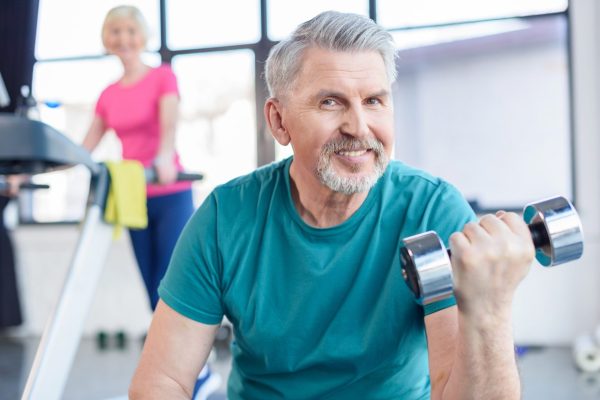 senior-sportsman-sitting-and-training-with-dumbbell-sportswoman-on-treadmill-behind-senior-fitness.jpg